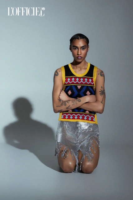 Fashion photo of a person kneeling with arms crossed, wearing a patterned top and silver skirt in a studio with grey background.