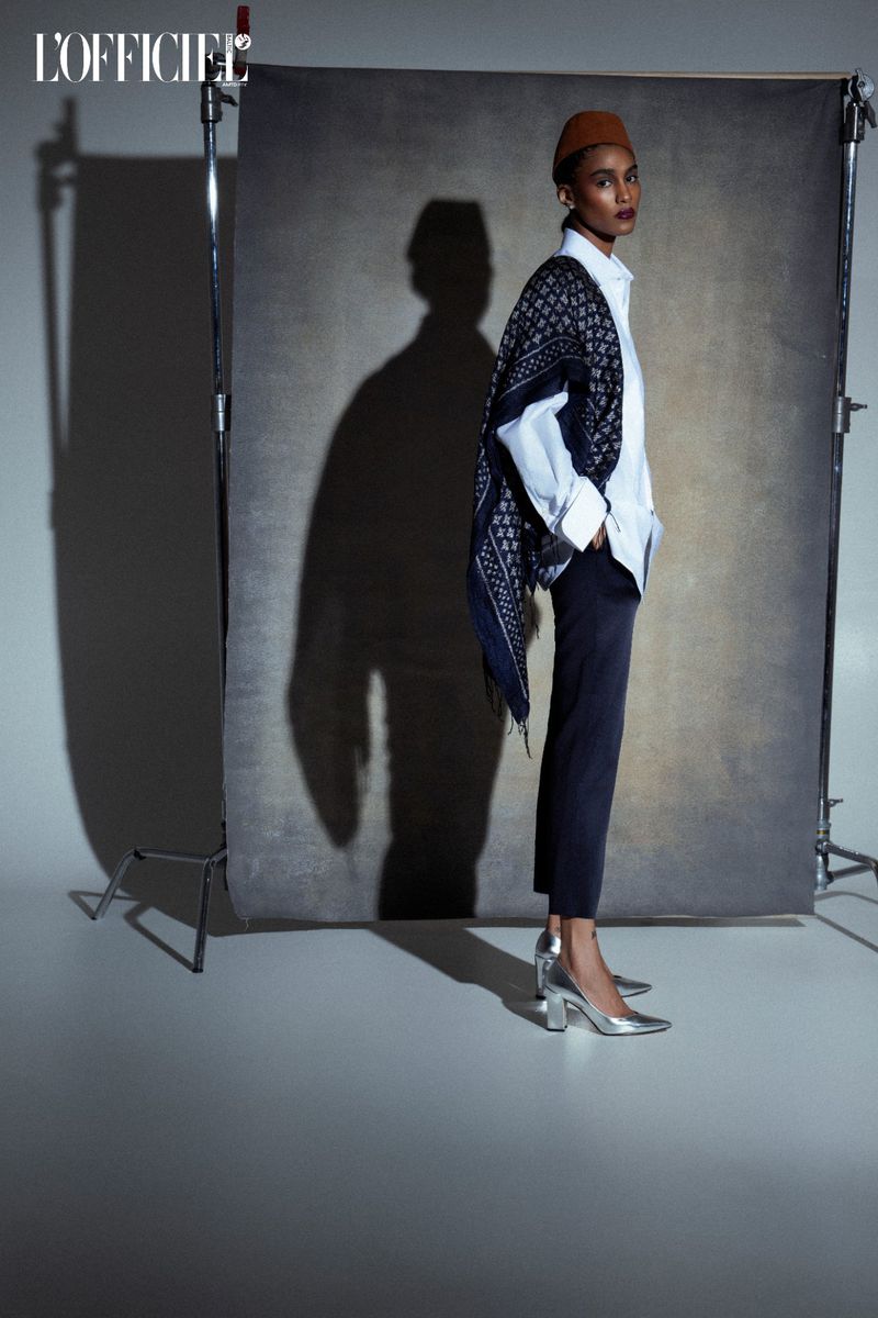 Fashion editorial shot of a dark-skinned model in a white shirt and brown hat posing against a gray backdrop in silver heels.