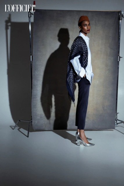 Fashion editorial shot of a dark-skinned model in a white shirt and brown hat posing against a gray backdrop in silver heels.