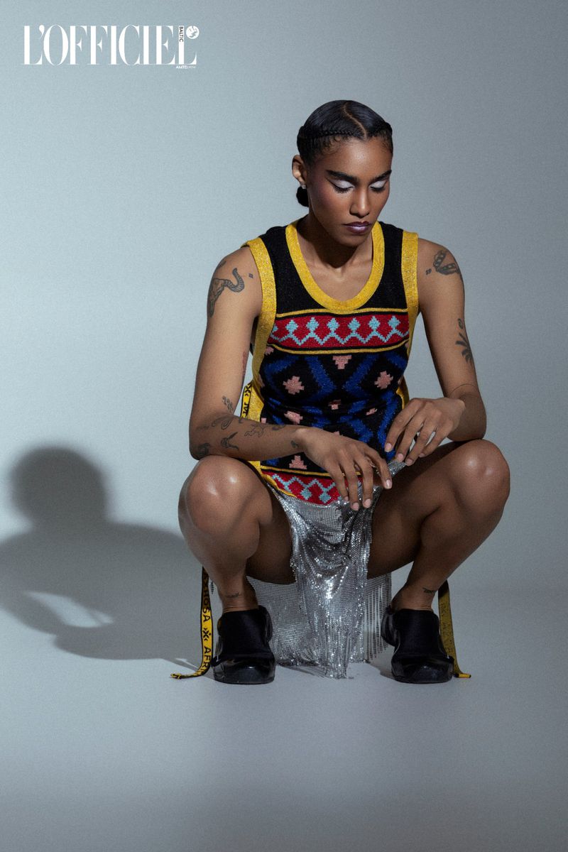 Woman in patterned dress and black boots squats in studio setting. Fashion editorial portrait with bright silver eye makeup.