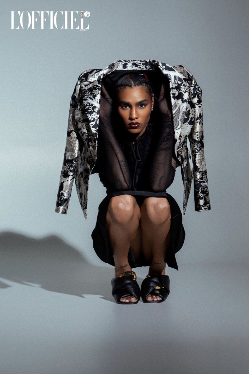 Fashion editorial shot of a Black adult female model squatting in a black dress and black slides, wearing a silver patterned jacket.