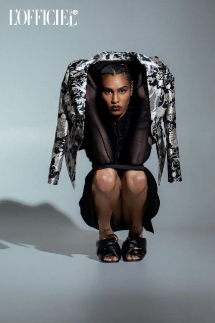 Fashion editorial shot of a Black adult female model squatting in a black dress and black slides, wearing a silver patterned jacket.