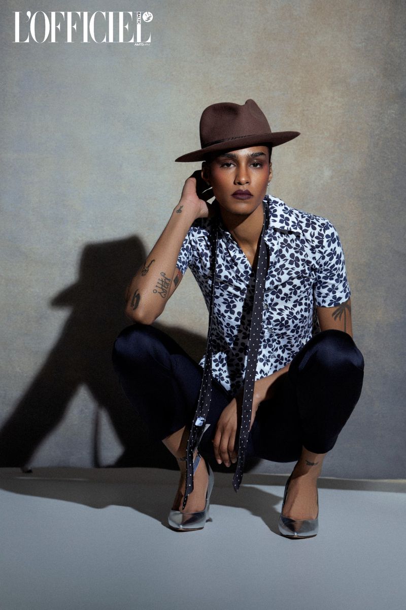 Person squats wearing a brown hat, flower-print shirt, black pants, and silver heels in a fashion photoshoot indoors.