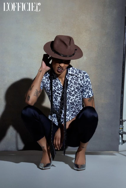 Adult male in floral shirt, dress pants, and pumps crouching in a photo studio, looking at the camera with a brown cowboy hat on.