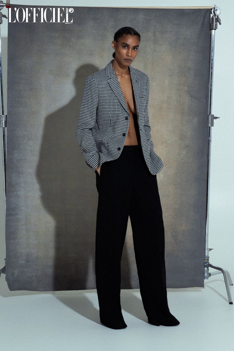 A woman in a houndstooth blazer and black pants poses against a grey backdrop in a studio, showcasing a high-fashion editorial look.