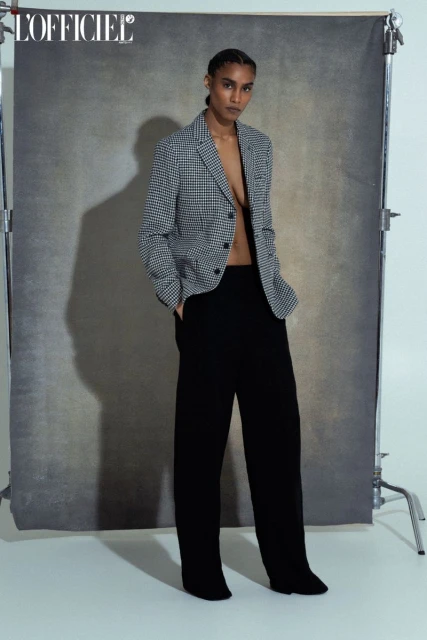 A woman in a houndstooth blazer and black pants poses against a grey backdrop in a studio, showcasing a high-fashion editorial look.