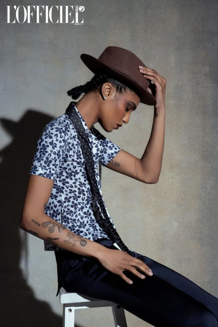 Adult female model in fashion shot wearing floral-print shirt, dark slacks, and hat in a studio setting with soft lighting.