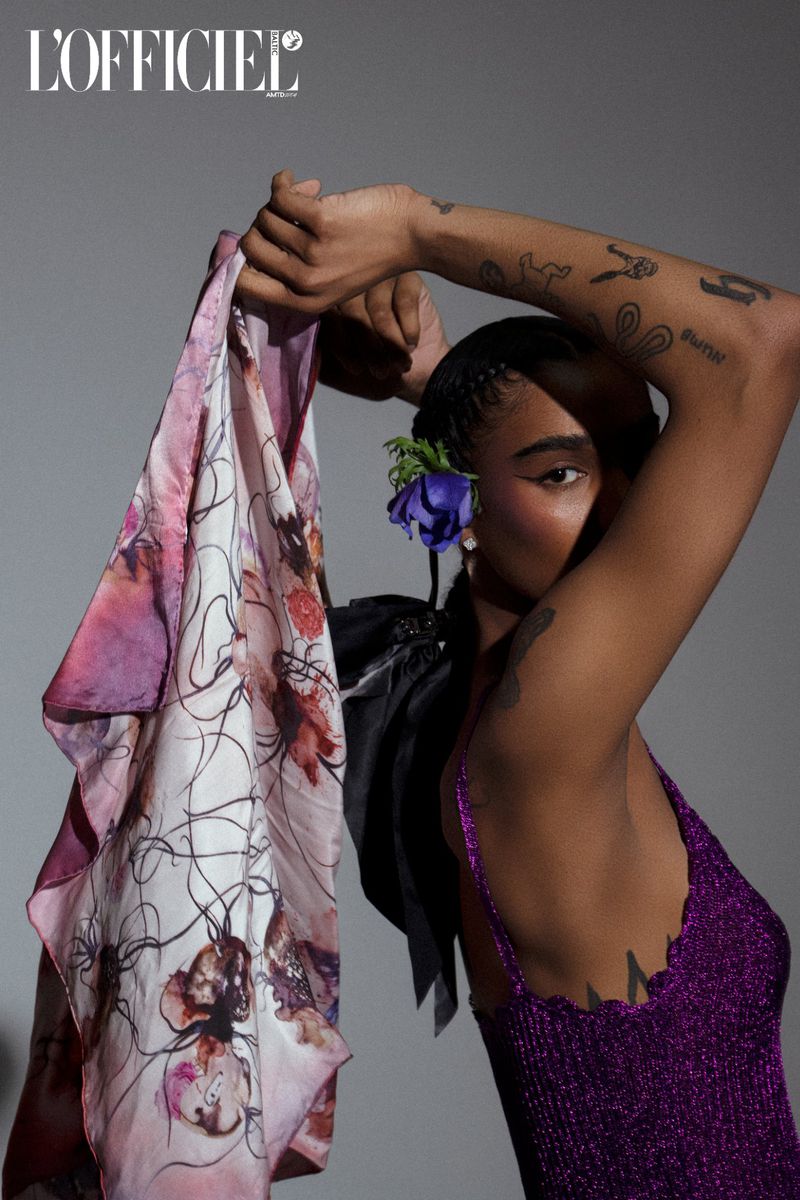 Portrait of a woman in a purple top with tattoos, floral scarf, and blue flower, against a grey backdrop and logo.