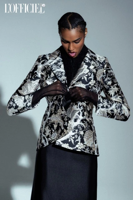 Fashionable Black model poses in a patterned blazer and skirt, accessorized with sheer gloves, against a neutral studio backdrop.