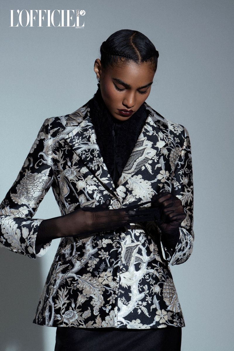 Fashion editorial shot of a Black woman wearing a black and white floral blazer, black top, and black gloves posing indoors.