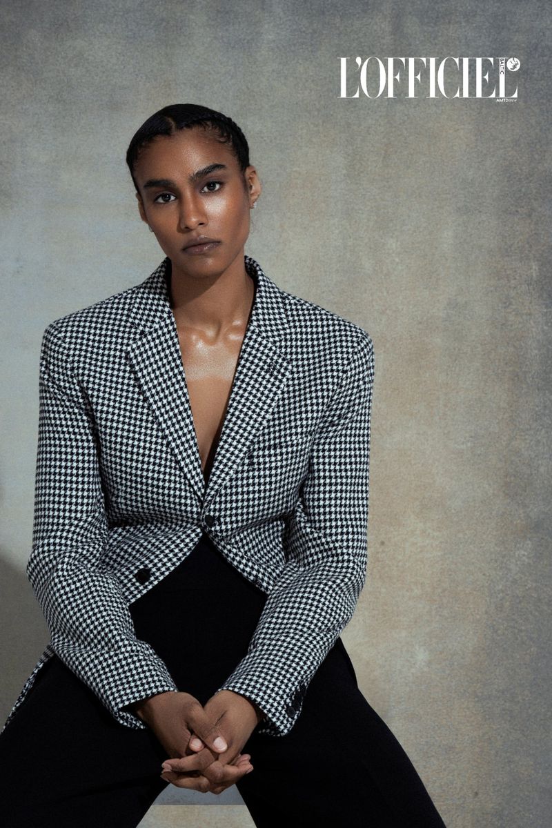 Adult woman in a houndstooth jacket poses indoors in front of a textured background. The fashion shot features the L'Officiel logo.