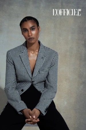 Adult woman in a houndstooth jacket poses indoors in front of a textured background. The fashion shot features the L'Officiel logo.