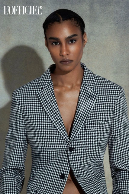 Adult female fashion model poses in a black and white houndstooth blazer looking directly at the camera in a studio setting.