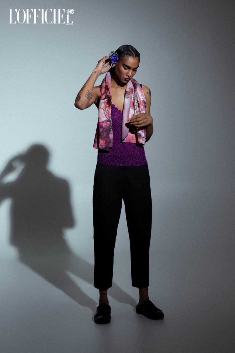 Fashion shot of androgynous person with blue flower in hair wearing scarf, purple top and black pants in front of grey background.