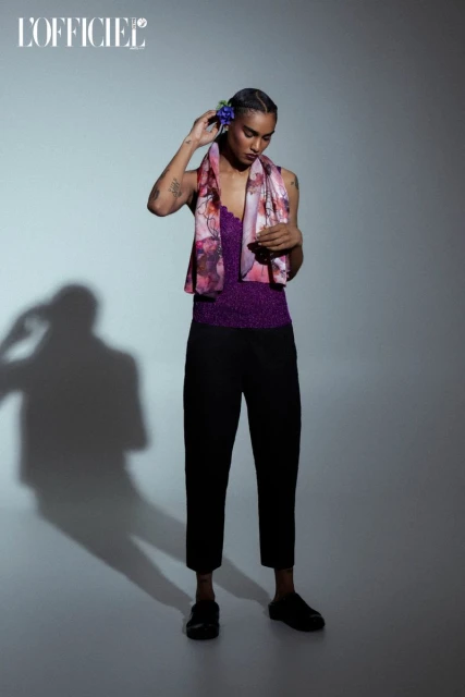 Fashion shot of androgynous person with blue flower in hair wearing scarf, purple top and black pants in front of grey background.