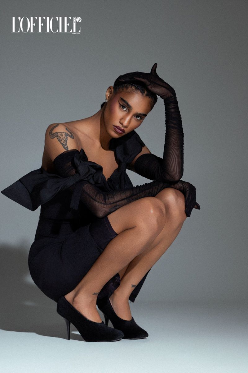 Fashionable woman in black dress and sheer gloves poses in photo studio. The model kneels wearing heels with ‘L'Officiel’ logo at top.