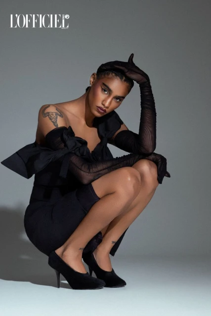 Fashionable woman in black dress and sheer gloves poses in photo studio. The model kneels wearing heels with ‘L'Officiel’ logo at top.