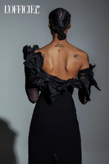 Fashion portrait from behind of a woman in a black dress with braided hair, tattoos, and ruffles against a gradient grey background.