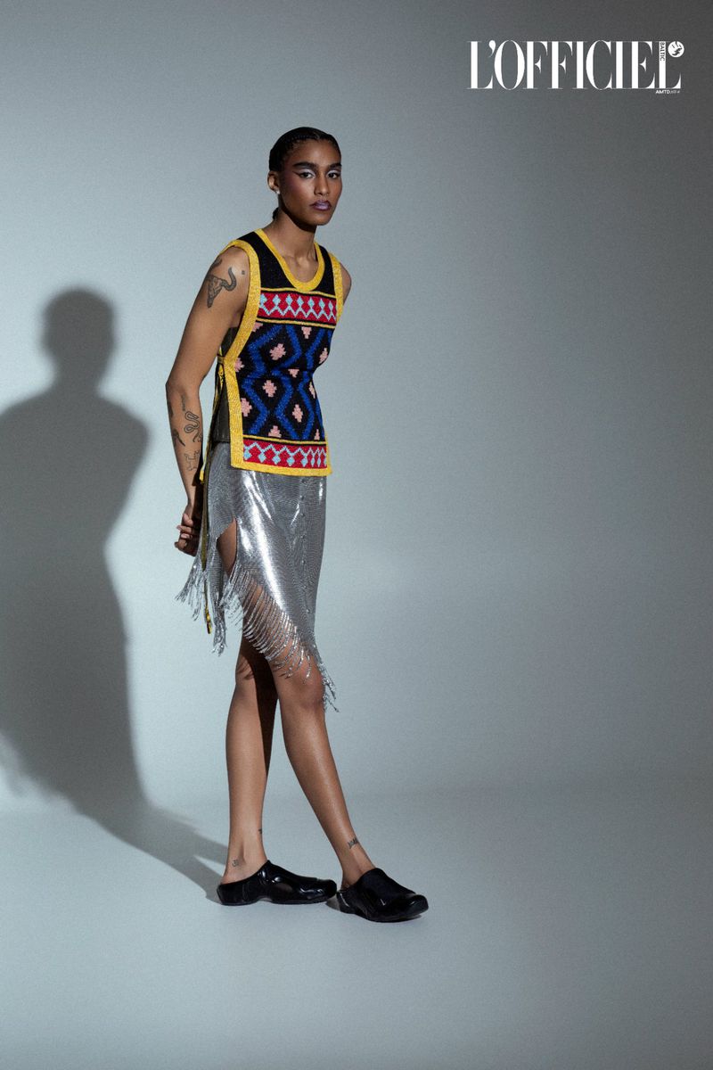 Woman poses in patterned top and silver skirt in a fashion studio. She wears black clogs against a gray background with studio lighting.