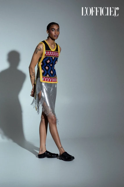 Woman poses in patterned top and silver skirt in a fashion studio. She wears black clogs against a gray background with studio lighting.