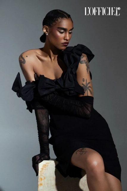 Elegant woman poses in a black dress with sheer gloves sitting against a gray backdrop with L'Officiel logo in the upper right.