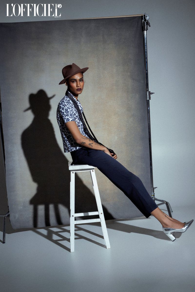 Model wearing a brown fedora and patterned top seated on a white stool against a mottled studio backdrop with a prominent shadow cast.