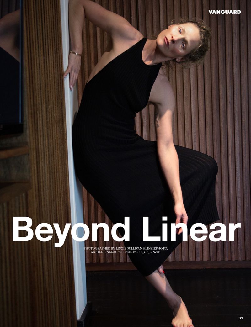 A slender woman in a black dress poses gracefully against a wooden wall, captured in a medium shot with soft, natural light indoors.