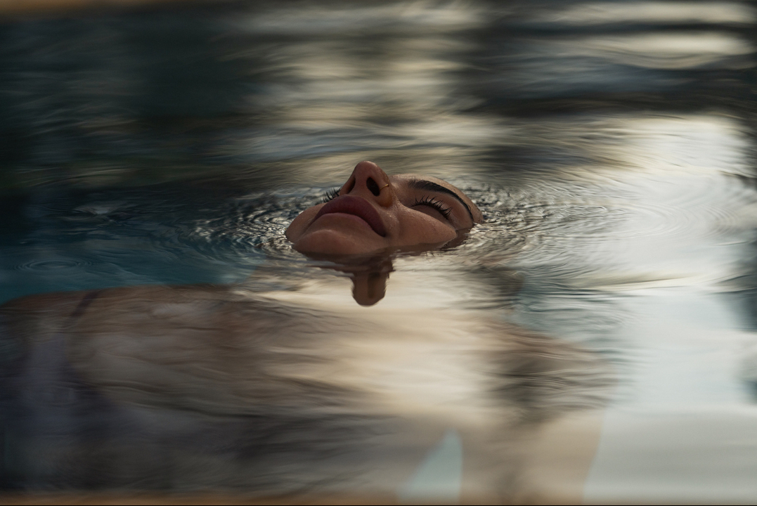 Close-up of a woman's face partially submerged in a pool of water, creating ripples and reflections in soft, natural lighting.