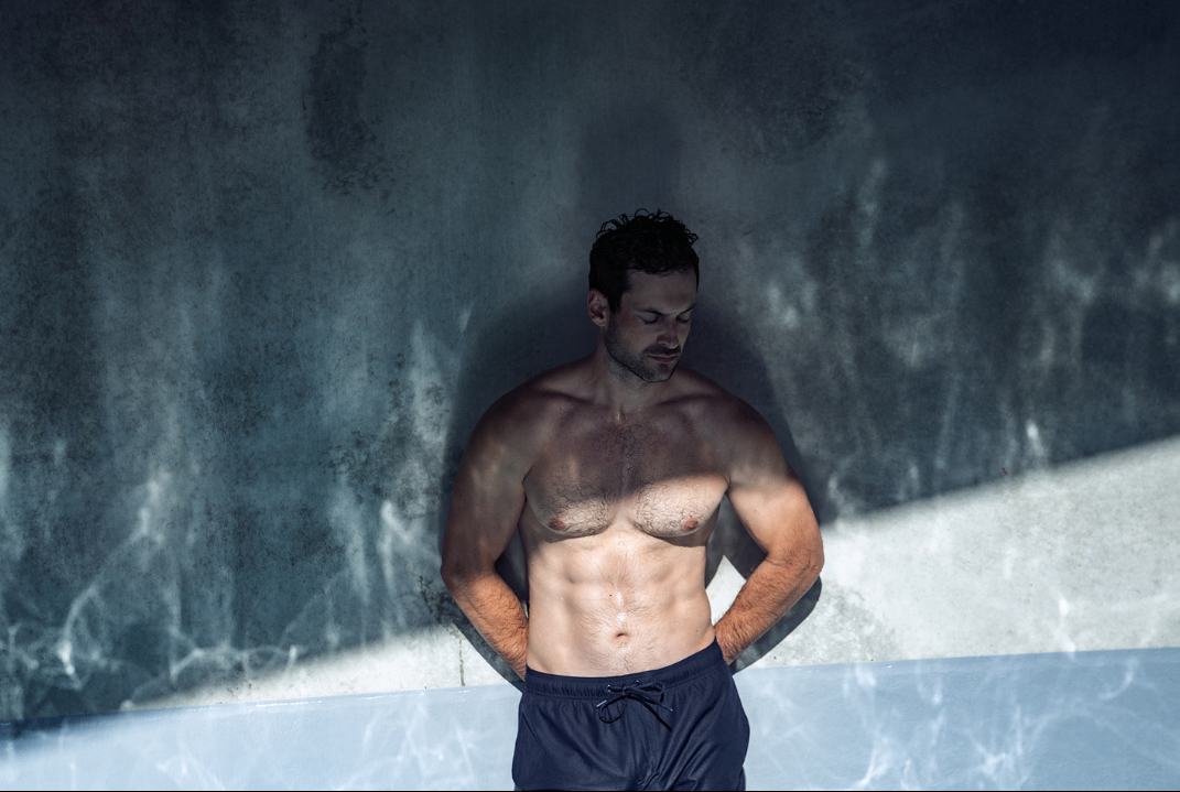 Athletic adult male with hands on hips poses in front of water-reflected wall wearing black shorts, lit with hard light and shadows.