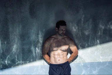 Shirtless man wearing dark swimming trunks leaning against a textured grey wall with water caustics light patterns.
