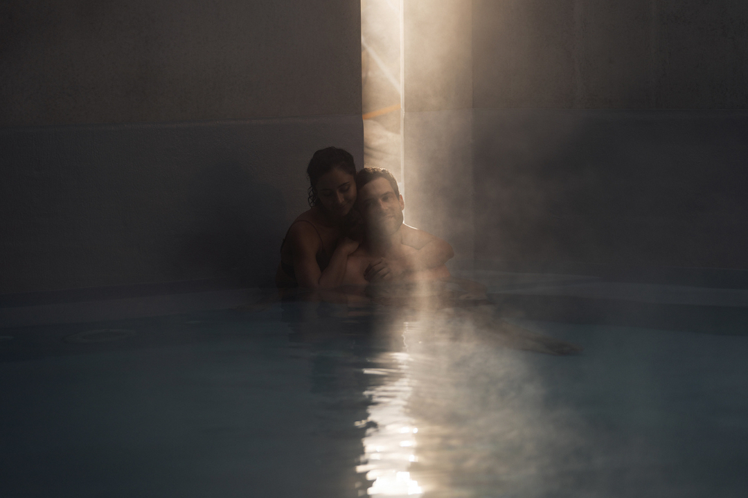 Couple embrace in a dimly lit indoor pool, a single ray of light highlighting the water's surface, creating a tranquil, intimate scene.