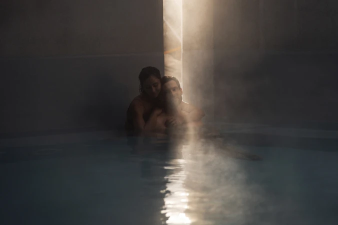 Couple embrace in a dimly lit indoor pool, a single ray of light highlighting the water's surface, creating a tranquil, intimate scene.