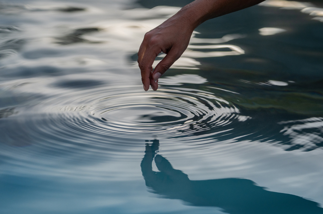 Close-up of an adult hand touching the surface of water creating a ripple effect on a body of water in an outdoor environment.