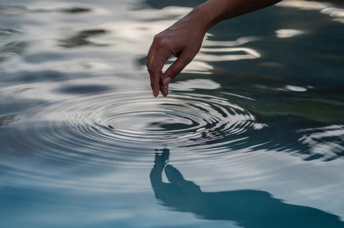 Close-up of an adult hand touching the surface of water creating a ripple effect on a body of water in an outdoor environment.