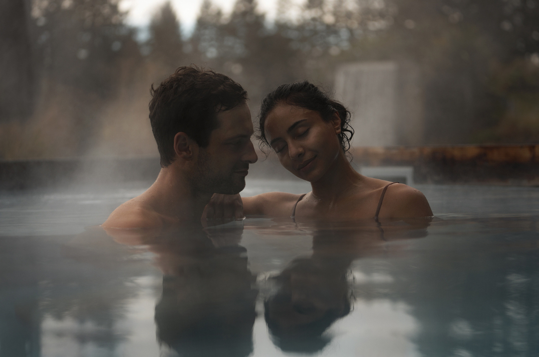 A couple is relaxing in a pool, with steam rising around them. The man and woman are partially submerged in the water, with greenery in the background.