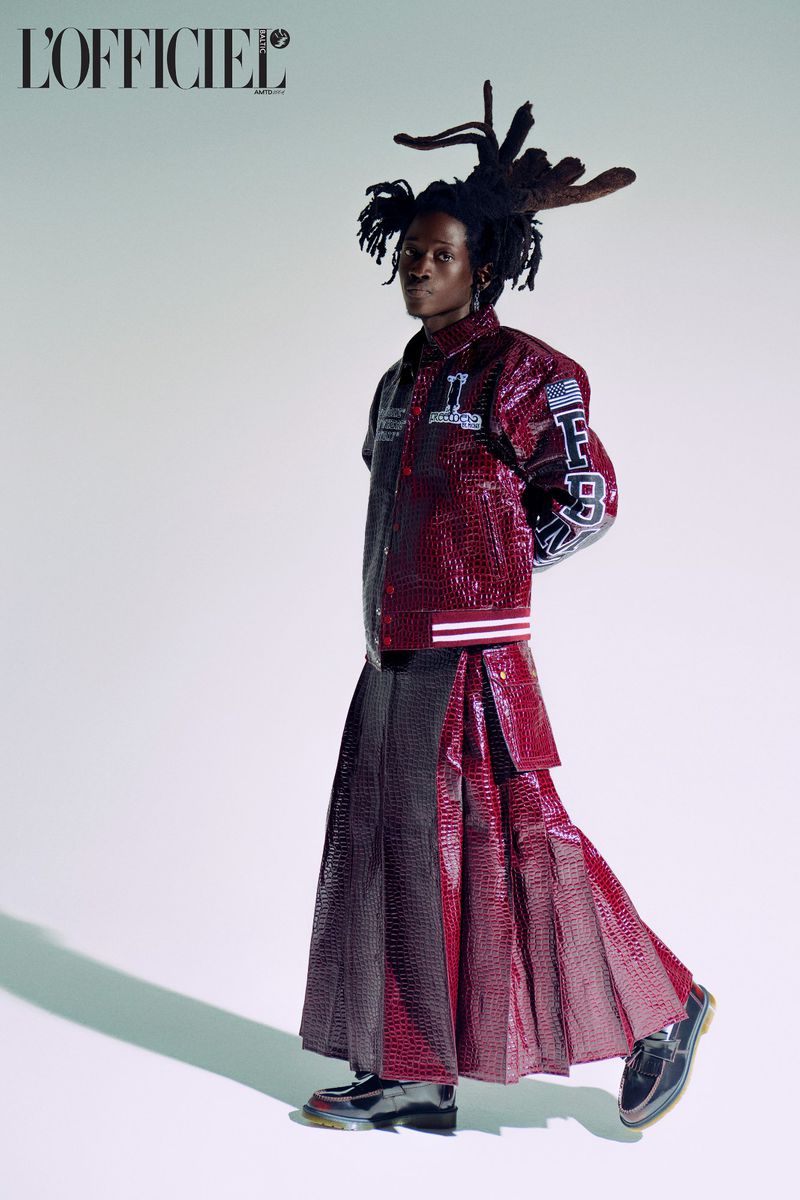 Fashion editorial shot of a Black model in a burgundy jacket and long skirt posing in front of a white backdrop with the magazine logo.