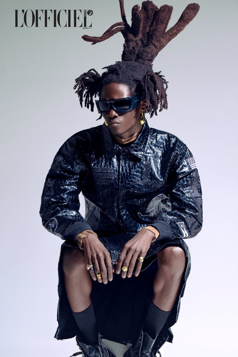 Fashion editorial shot of male model in black jacket and dreadlocks wearing sunglasses and jewelry sitting against grey studio backdrop.