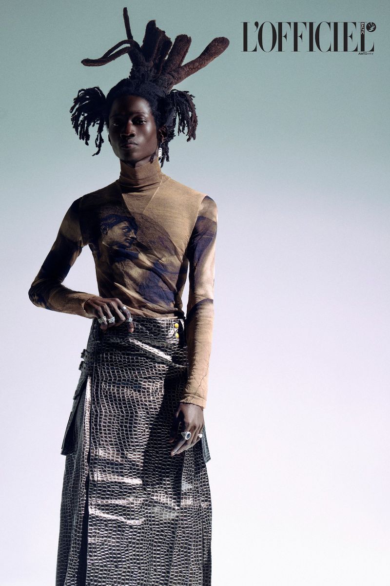 A person with dark skin and elaborate dreadlocks poses in a fashion shot, wearing a patterned top and skirt, against a light background.