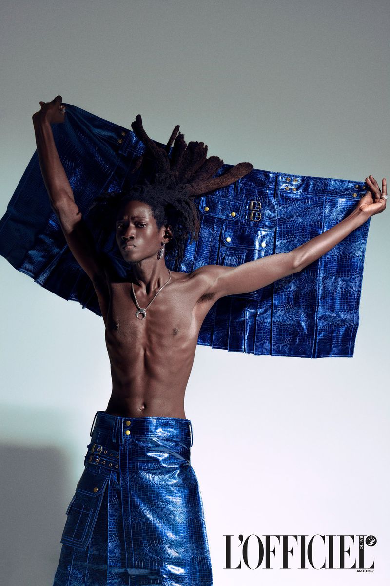 Fashion editorial shot of a slender, dark-skinned man modeling clothing with blue croc pattern. He wears skirt and holds up matching jacket.