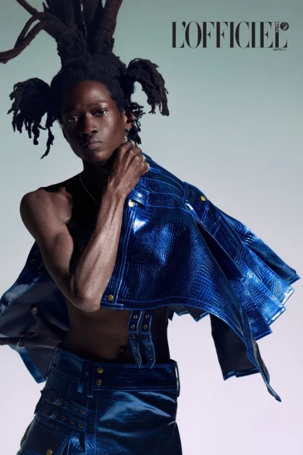 Dark-skinned person with dreadlocks wearing a blue reptile-skin jacket and skirt, posing for L'Officiel fashion editorial against a light background.