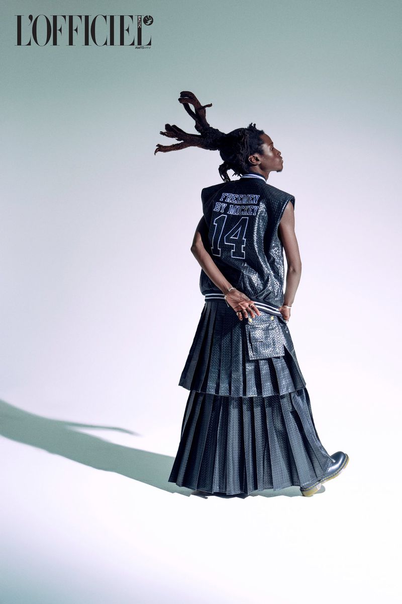 Back view of a slender person with dark skin, wearing a black sequin vest, long pleated skirt, and boots, against a white backdrop.