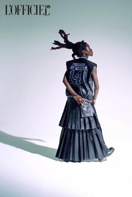 Back view of a slender person with dark skin, wearing a black sequin vest, long pleated skirt, and boots, against a white backdrop.