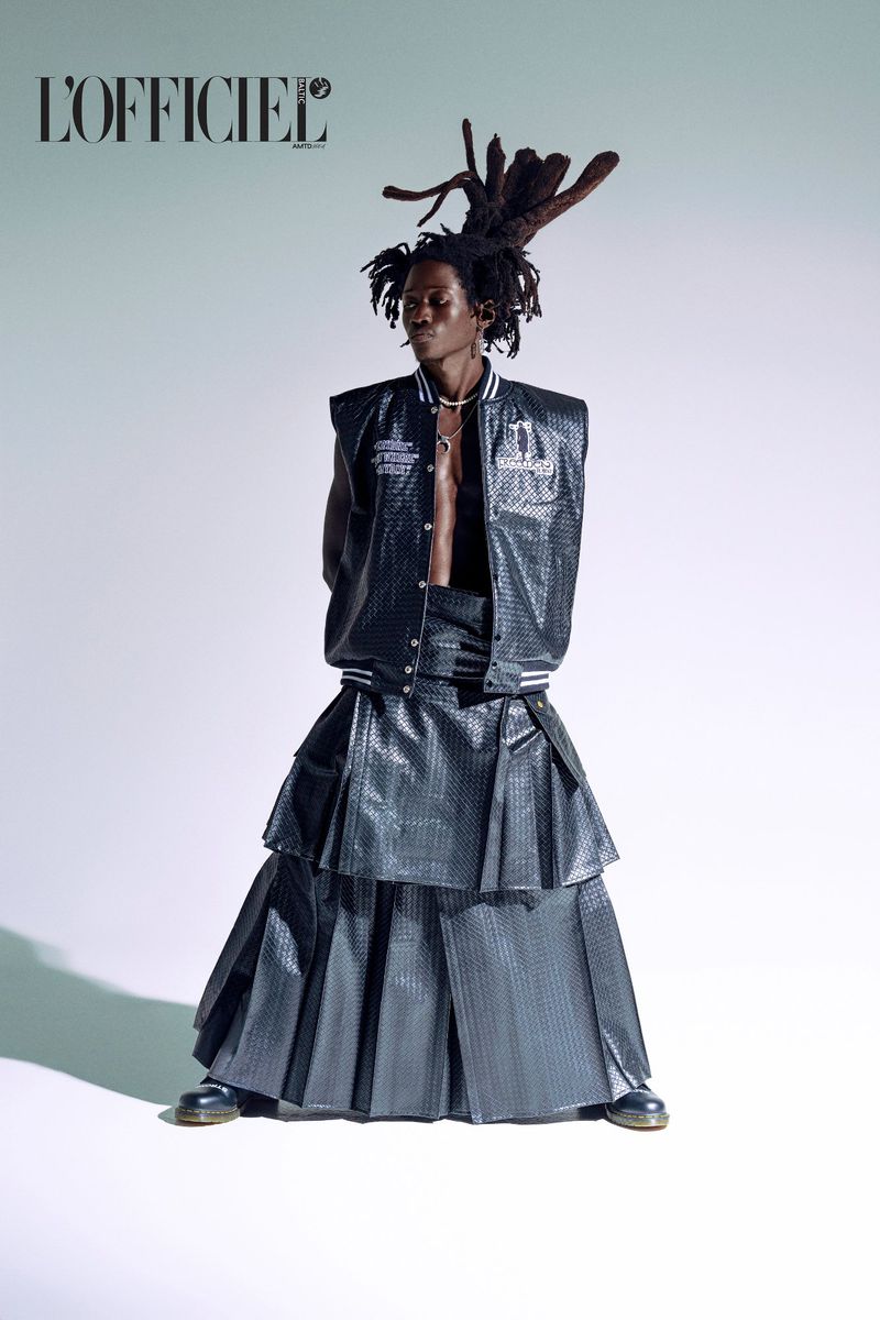 Editorial fashion shot of a male model in a layered black skirt and vest, posing in a studio with the L'Officiel magazine logo.