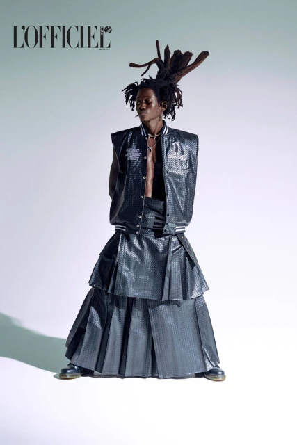 Editorial fashion shot of a male model in a layered black skirt and vest, posing in a studio with the L'Officiel magazine logo.