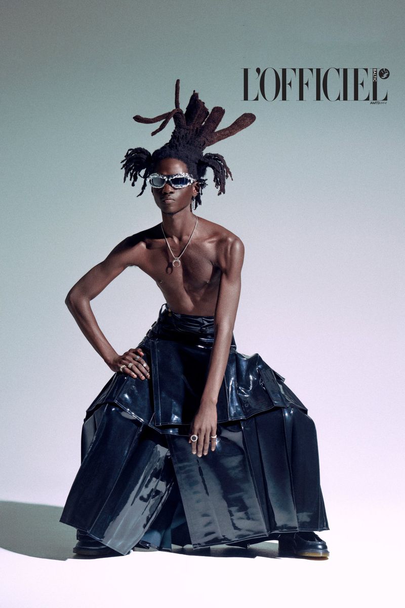 Fashion editorial shot of a bare-chested Black male model with an elaborate updo and silver sunglasses, wearing a shiny black skirt.
