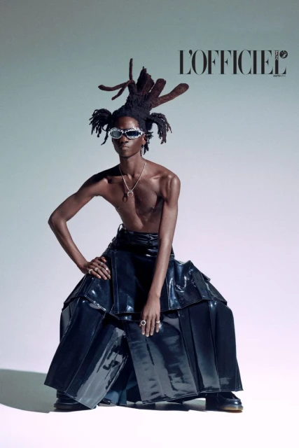 Fashion editorial shot of a bare-chested Black male model with an elaborate updo and silver sunglasses, wearing a shiny black skirt.