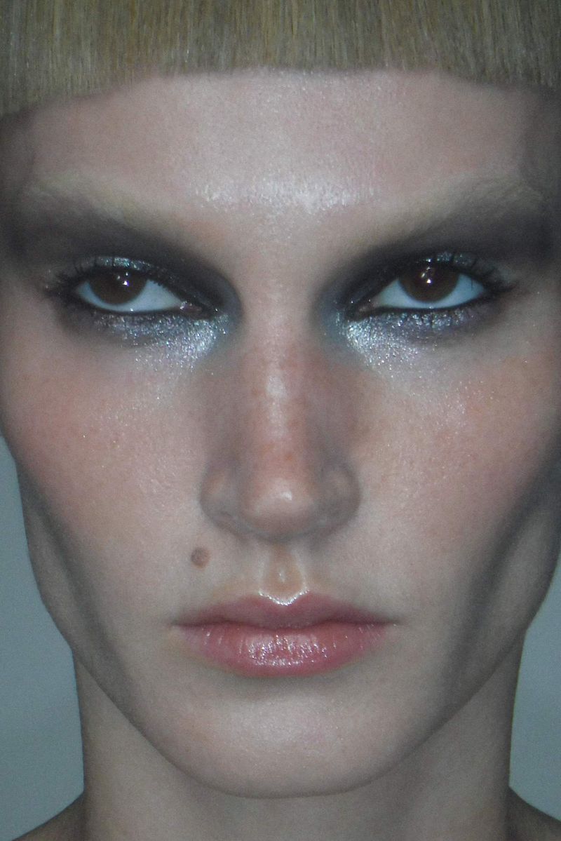 Close-up portrait of a blonde adult woman with heavy silver eyeshadow makeup, glossy lips, and a mole on her cheek, on a plain background.