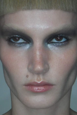 Close-up portrait of a blonde adult woman with heavy silver eyeshadow makeup, glossy lips, and a mole on her cheek, on a plain background.