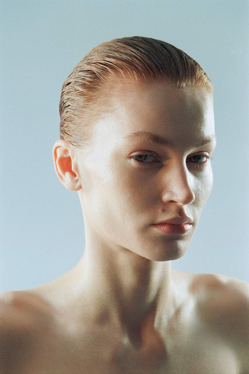 Close-up portrait of a fair-skinned young adult female with slicked-back auburn hair against a pale blue background in a studio setting.