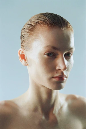 Close-up portrait of a fair-skinned young adult female with slicked-back auburn hair against a pale blue background in a studio setting.